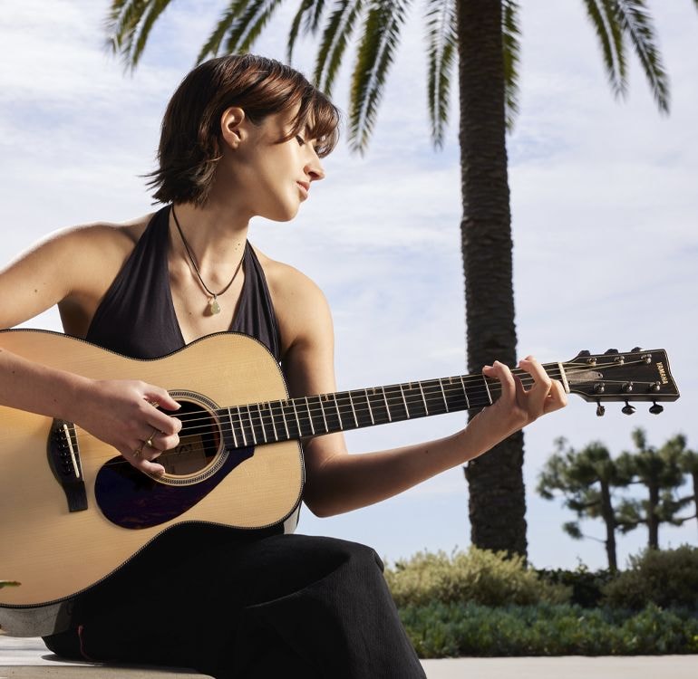 Guitarist Sophia James playing an FS9 guitar sits in front of a palm tree outside and plays an FS9 acoustic guitar.